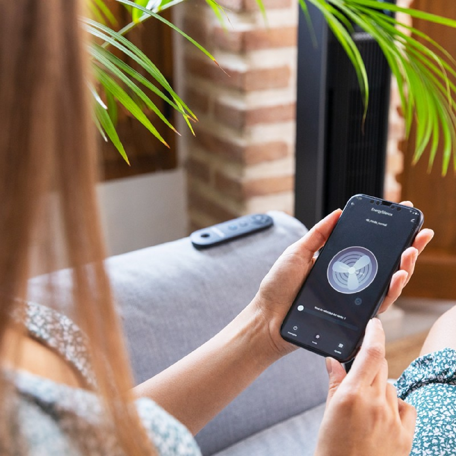 Kobieta używa smartfona z aplikacją do sterowania wentylatorem. Roślina i ceglana ściana w tle.