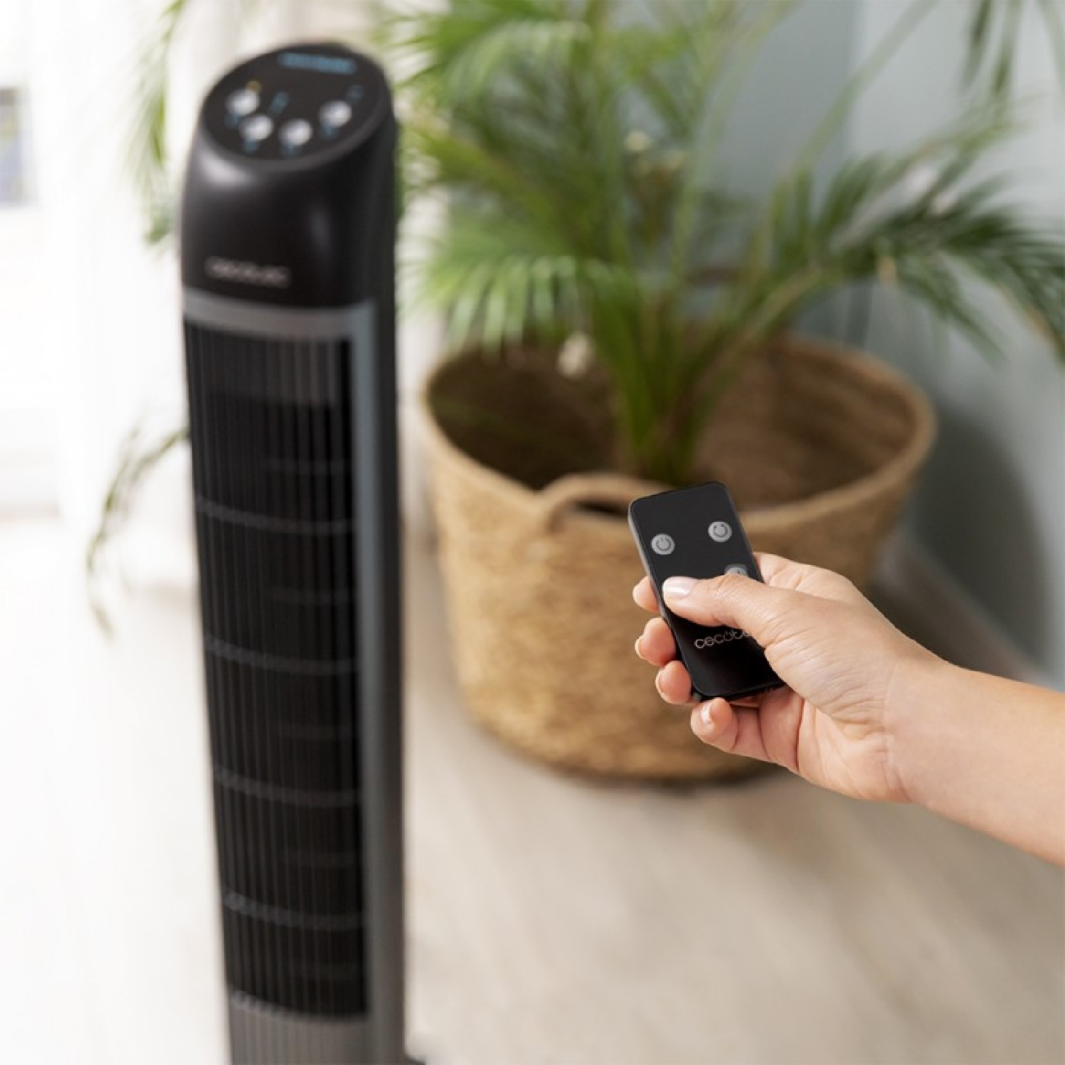 Czarny wentylator wieżowy z dłonią trzymającą pilota przed rośliną w wiklinowym koszu.