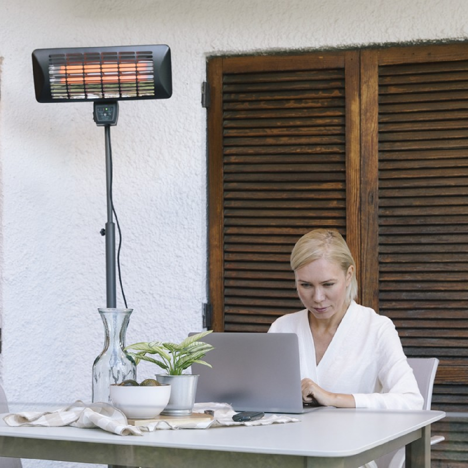 Kobieta używająca laptopa na zewnątrz przy grzejniku. Scena zawiera stół, rośliny i drewniane okiennice.