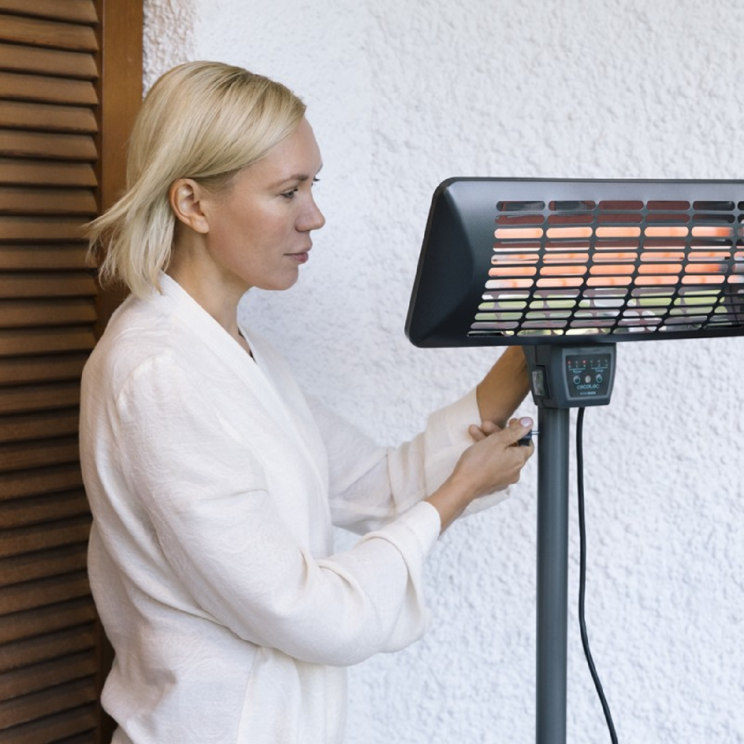 Kobieta reguluje czarny grzejnik zewnętrzny. Ma na sobie biały szlafrok. Tło jest białe.