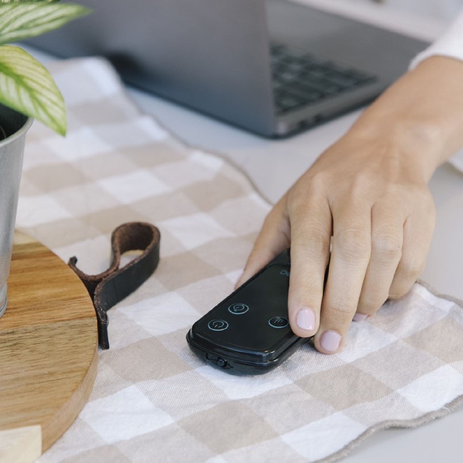 Ręka osoby trzymająca czarny pilot na kraciastej szmatce. W tle widoczny jest laptop.