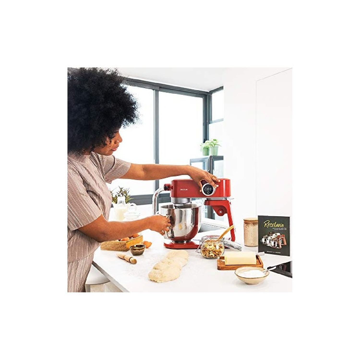 Donna usa un mixer rosso su un bancone bianco. Ingredienti e libro di cucina presenti.