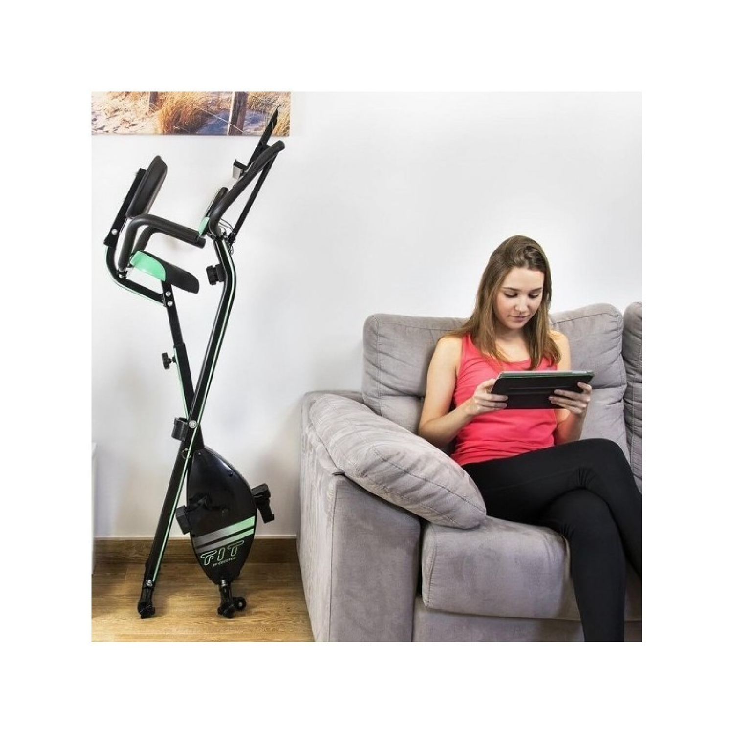 Donna su un divano che usa un tablet accanto a una cyclette. Parete bianca e opera d'arte sullo sfondo.