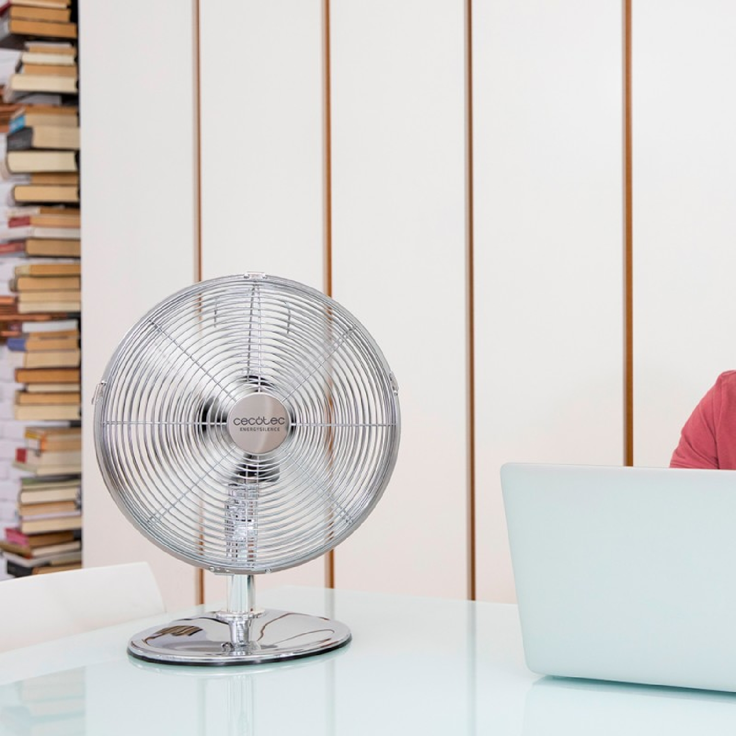 Ventilatore elettrico argentato su un tavolo bianco. Libri a sinistra e laptop a destra.