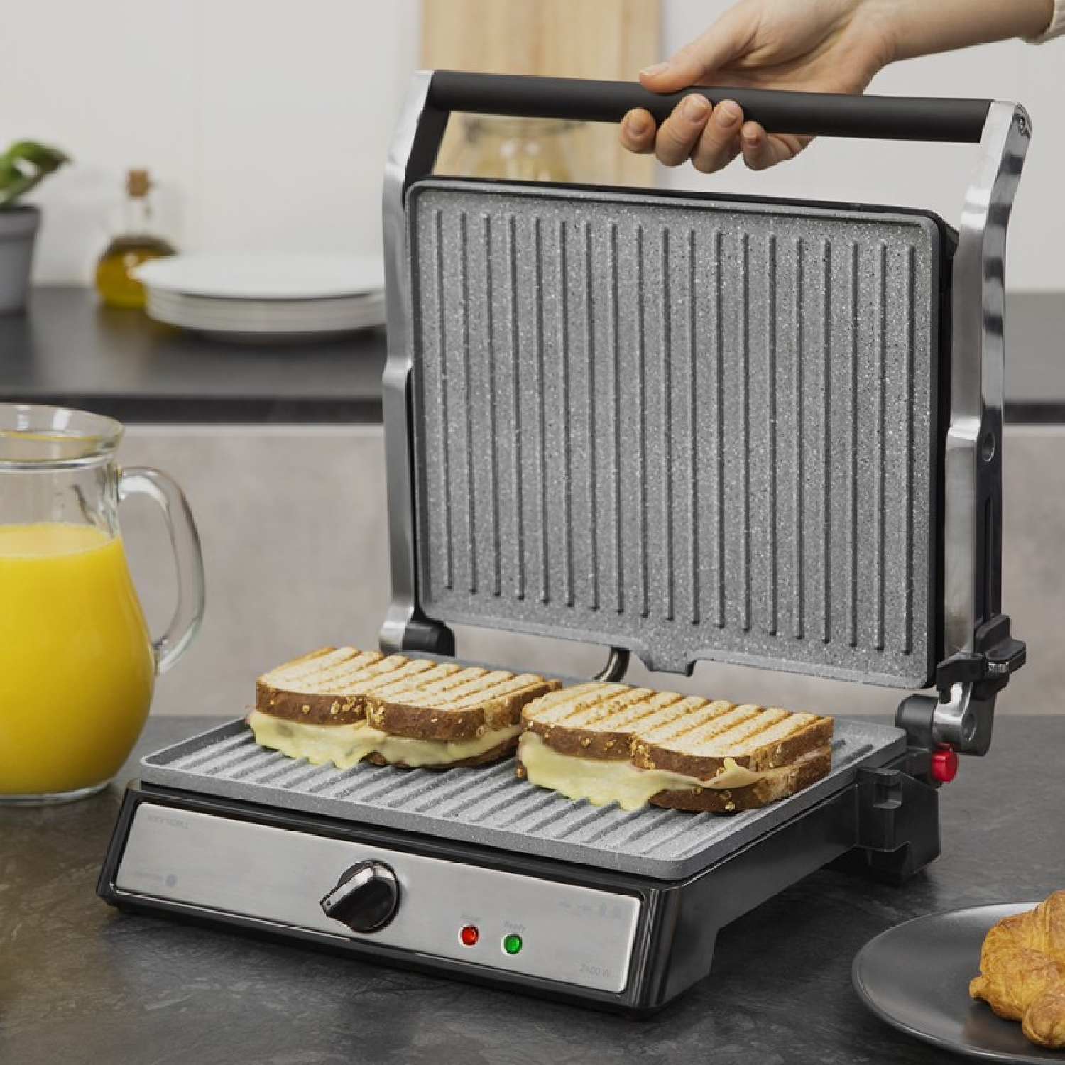 Ręka otwiera prasę do panini z dwoma kanapkami w środku, na blacie.