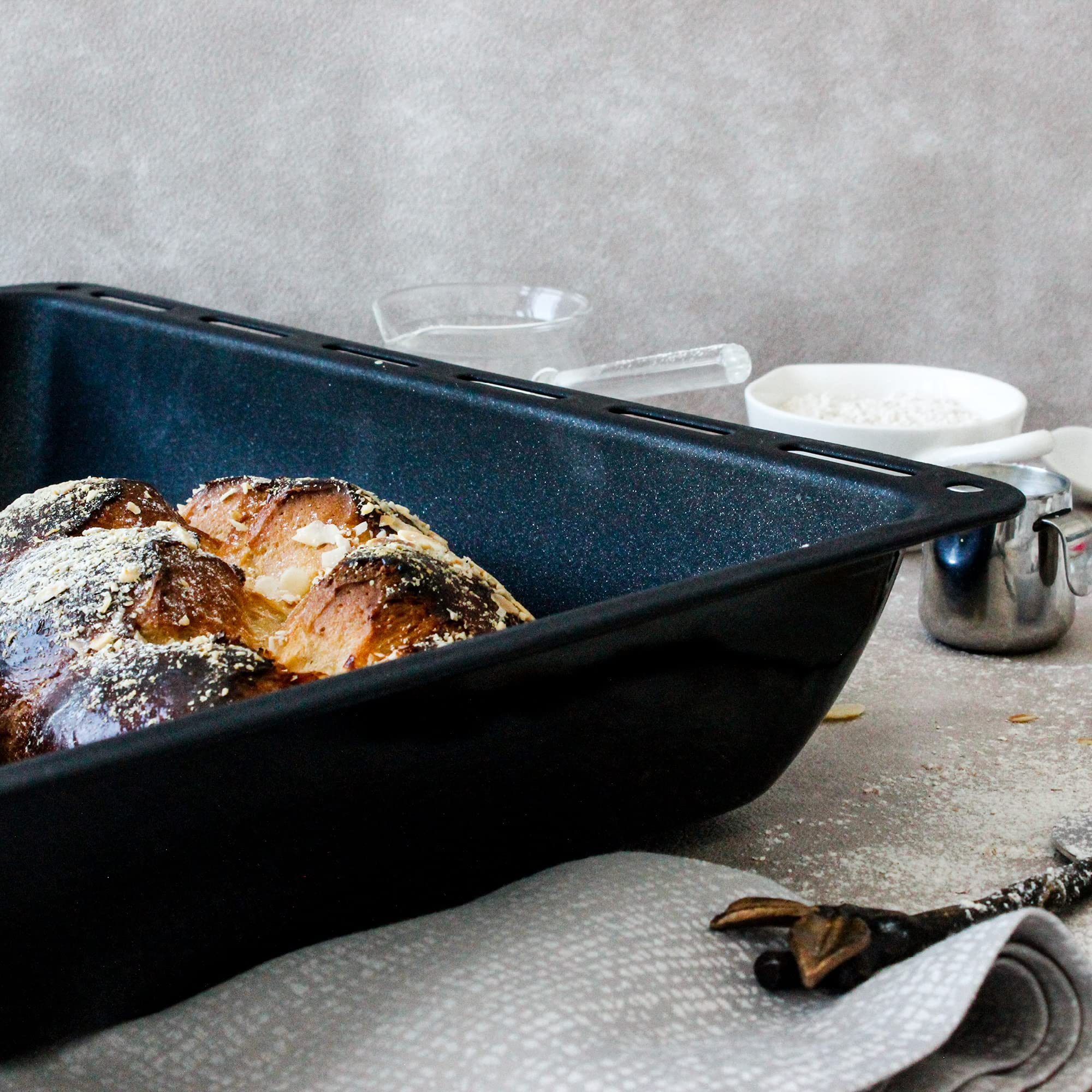 Schwarze Backform mit Brot. Mehl und Zutaten auf grauem Hintergrund sichtbar.