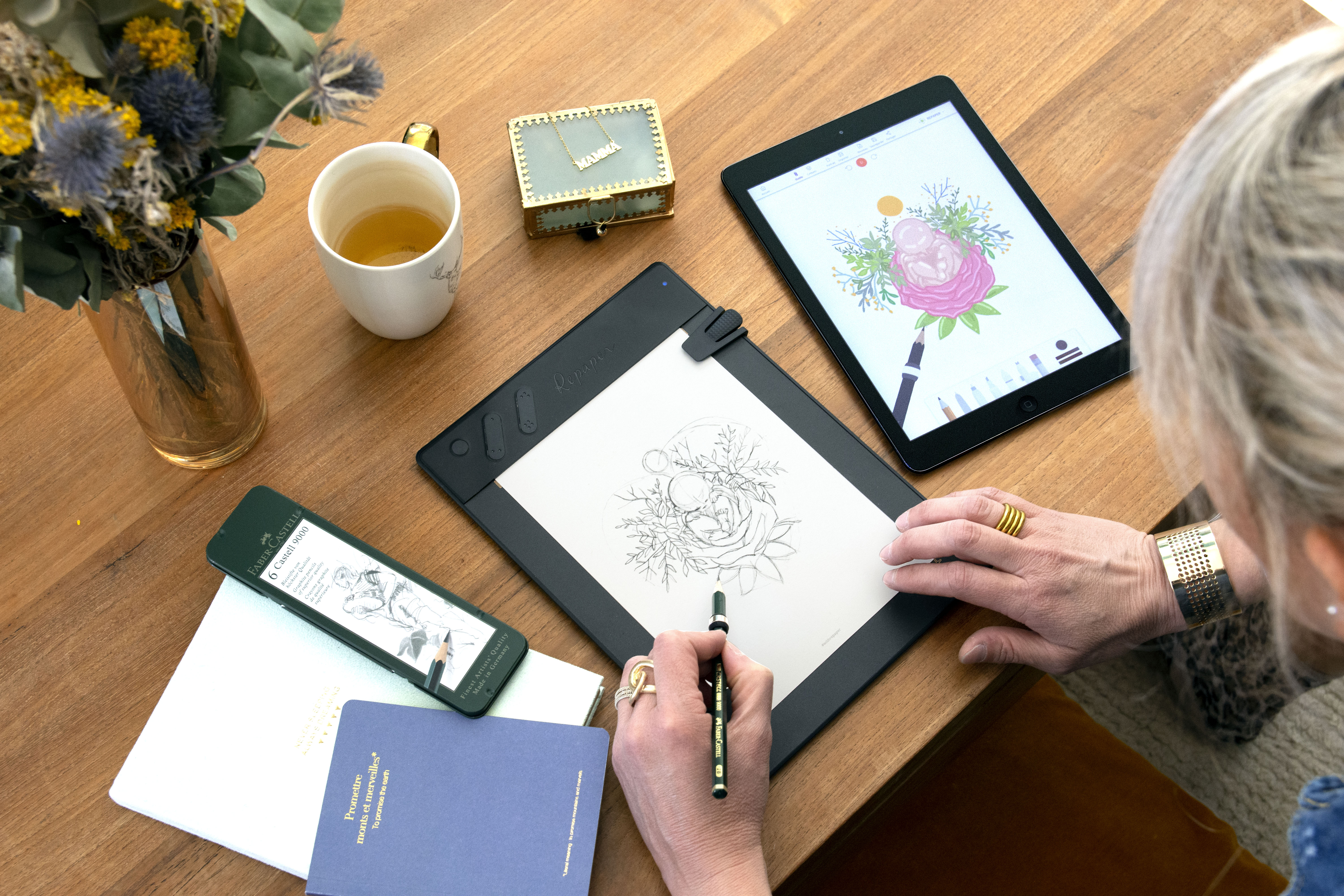 Person zeichnet auf einem Tablet, Notizbuch und einer Kaffeetasse auf einem Holztisch.
