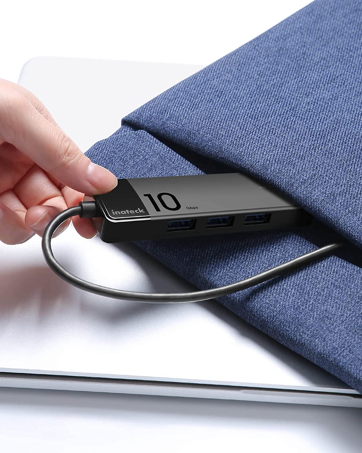 Una mano inserta un concentrador USB negro en una funda de tela azul. El concentrador tiene puertos USB y etiqueta '10 Gbps'.