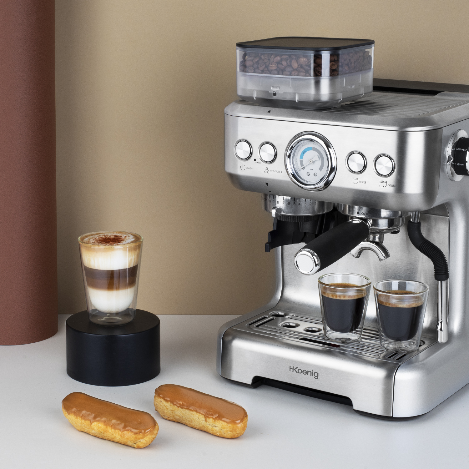 Macchina per espresso argentata, contenitore di chicchi di caffè, due shot di espresso e due éclair. Sfondo chiaro.