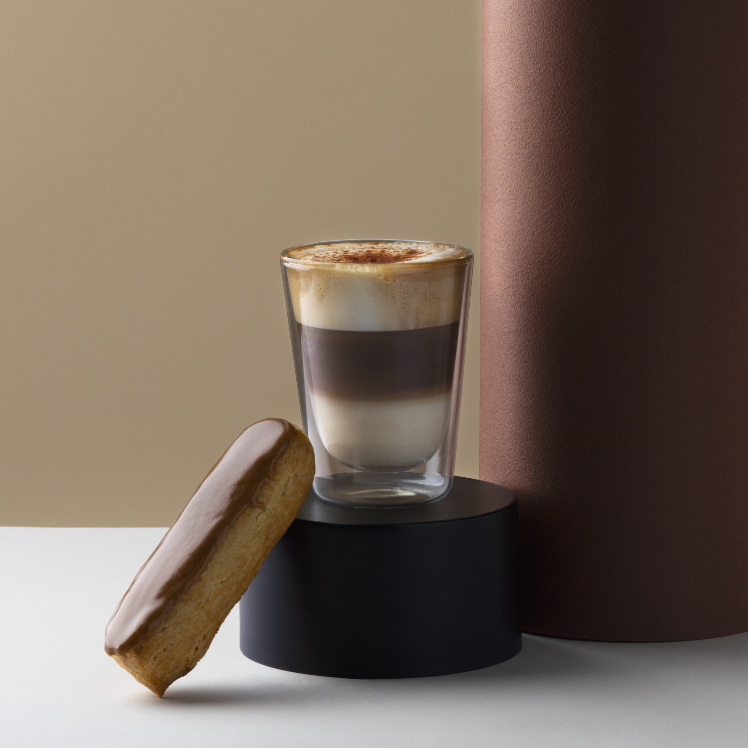 Un cappuccino e un éclair al caramello su un supporto nero con sfondo cilindrico marrone.