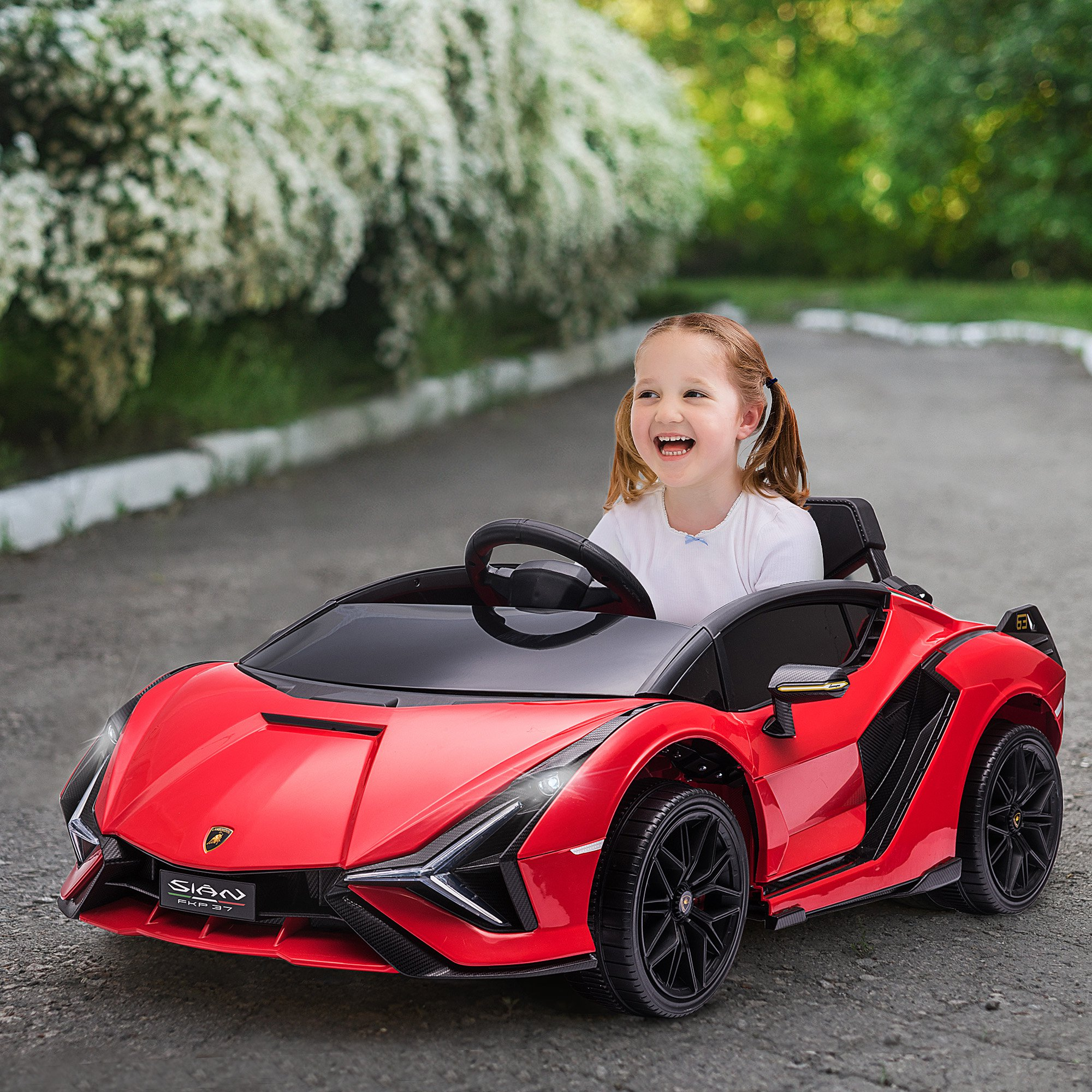 Un niño sonríe mientras conduce un coche de juguete rojo al aire libre. El coche es un modelo Lamborghini.