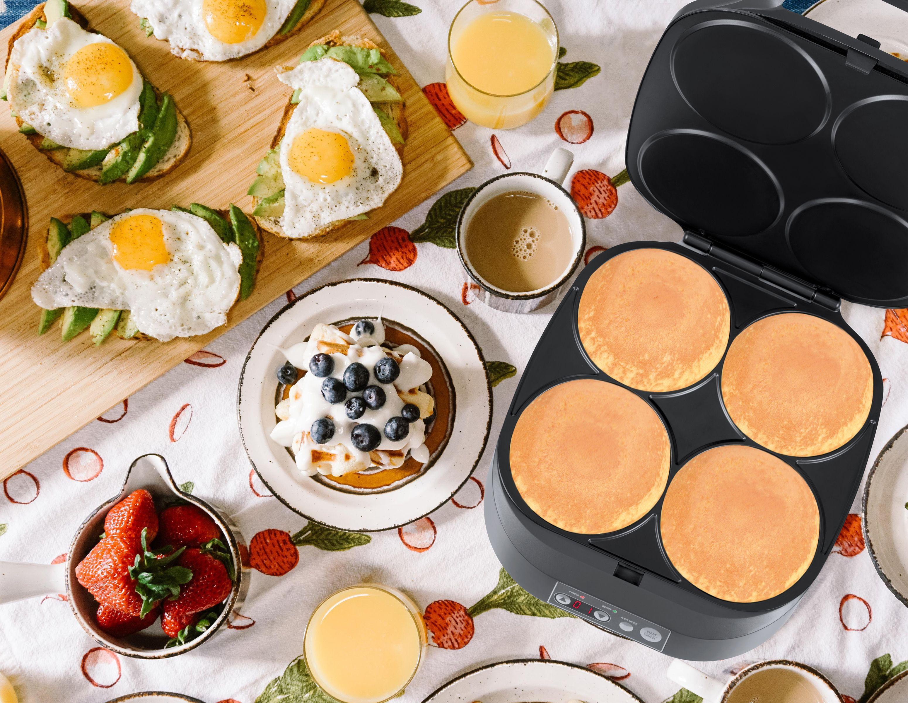 Mesa de desayuno con panqueques, huevos, tostadas de aguacate y jugo. A la derecha hay una máquina para hacer panqueques negra.