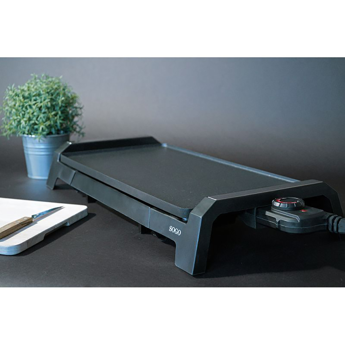 Plancha eléctrica negra con una pequeña planta y una tabla de cortar sobre una superficie oscura.