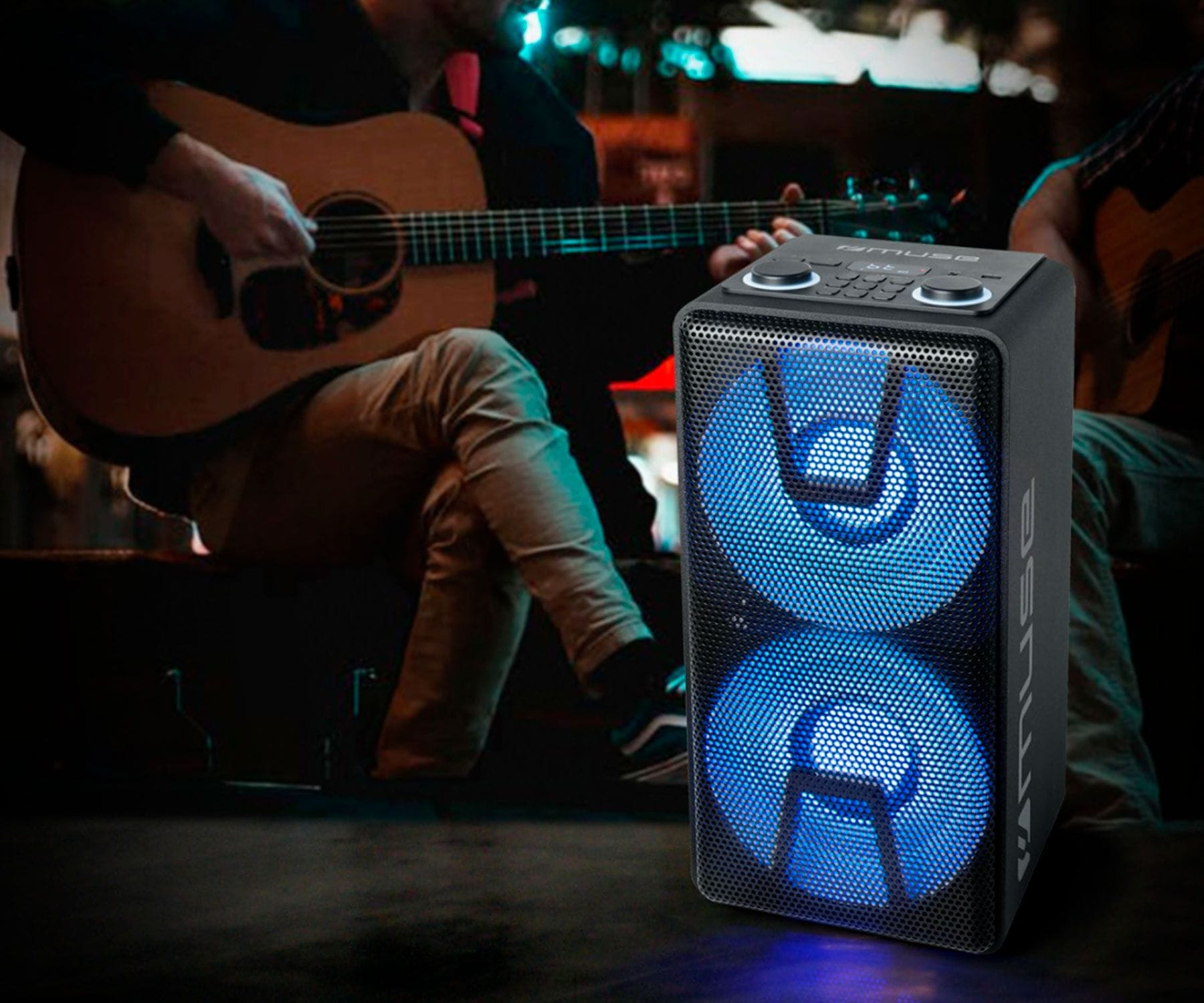 Czarny głośnik z niebieskimi światłami grający, ludzie grający na gitarze w tle, w pomieszczeniu.