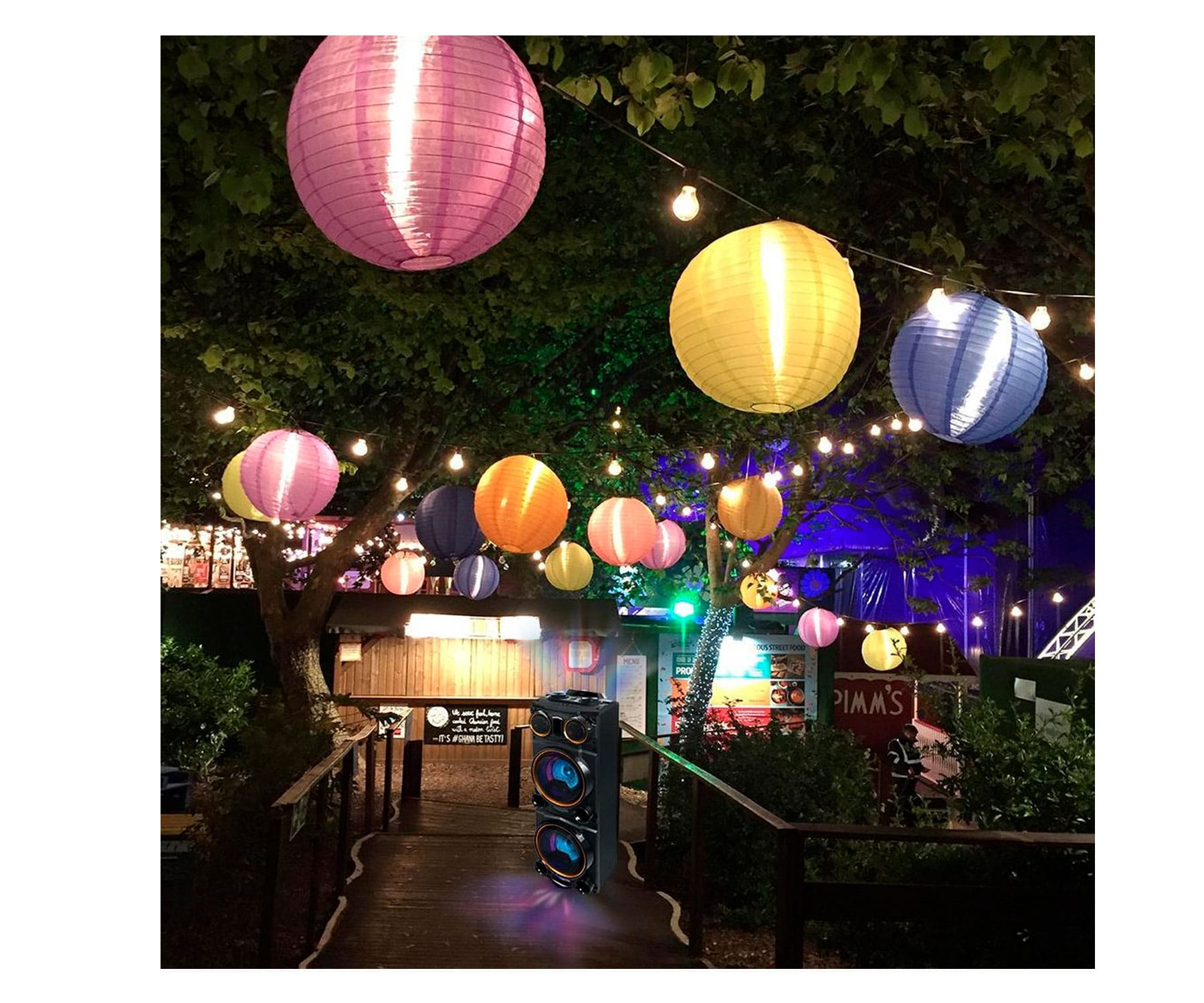 Scena na zewnątrz z kolorowymi lampionami, głośnikami i drewnianym mostem w nocy.