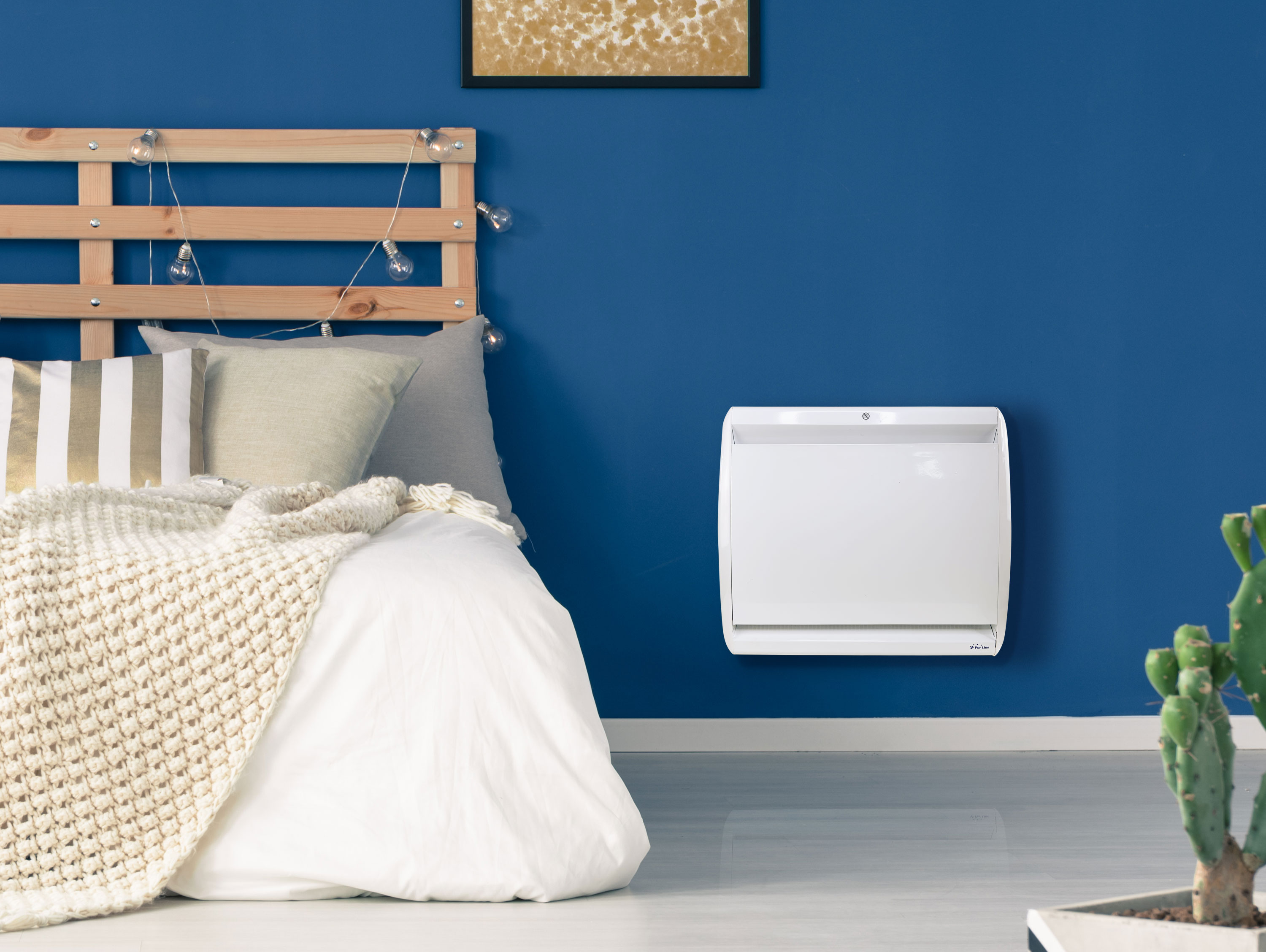 Weißer Wandheizer an blauer Wand in einem Schlafzimmer. Bett mit Decke und Kissen sichtbar. Ein Kaktus steht auf dem Boden.