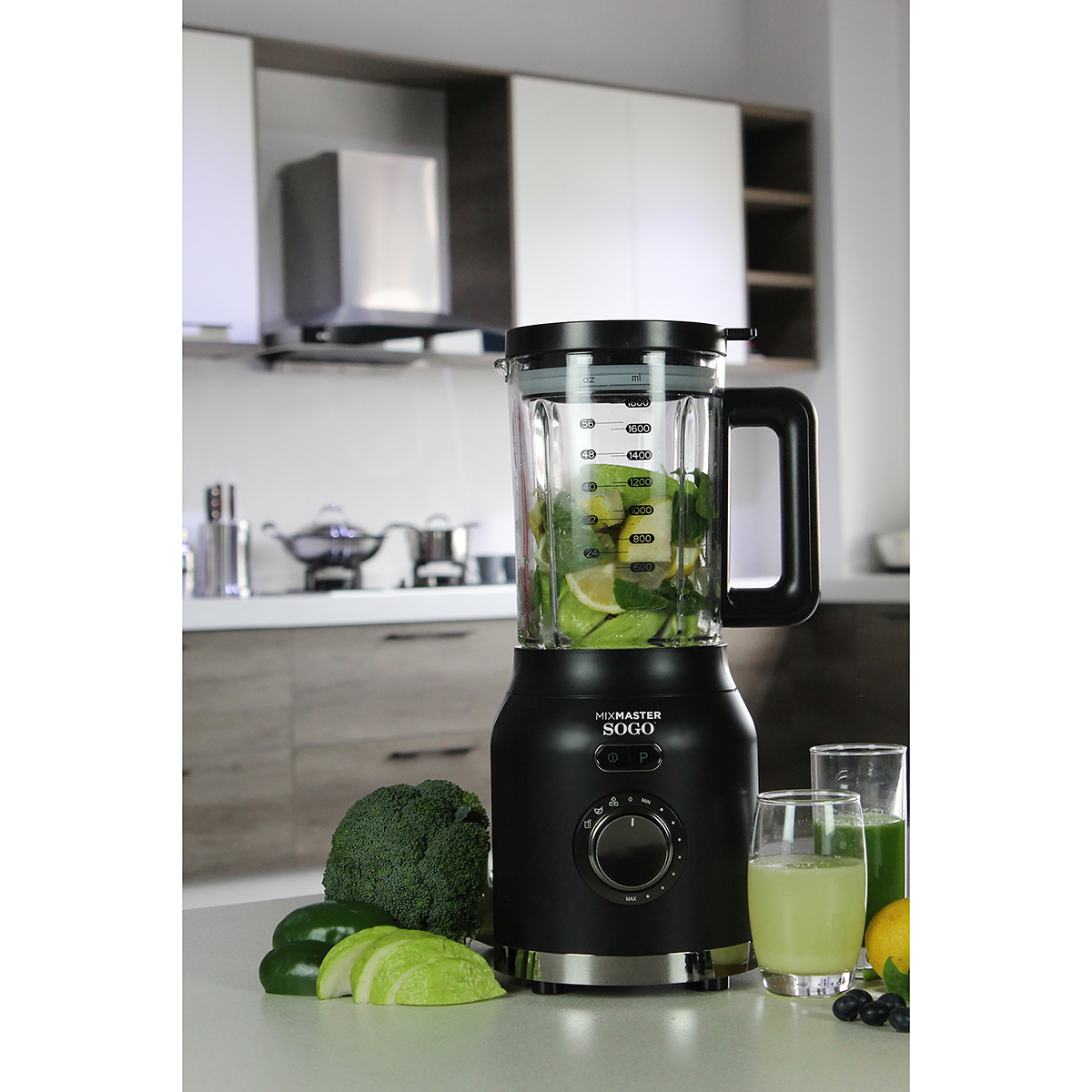 Frullatore nero con ingredienti dentro e intorno. Cucina sullo sfondo.