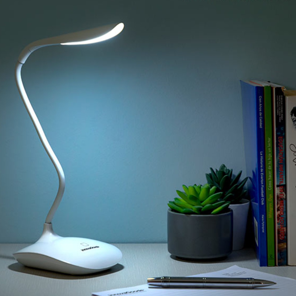 Lampada da scrivania bianca con collo flessibile su scrivania, libri e piante sullo sfondo.
