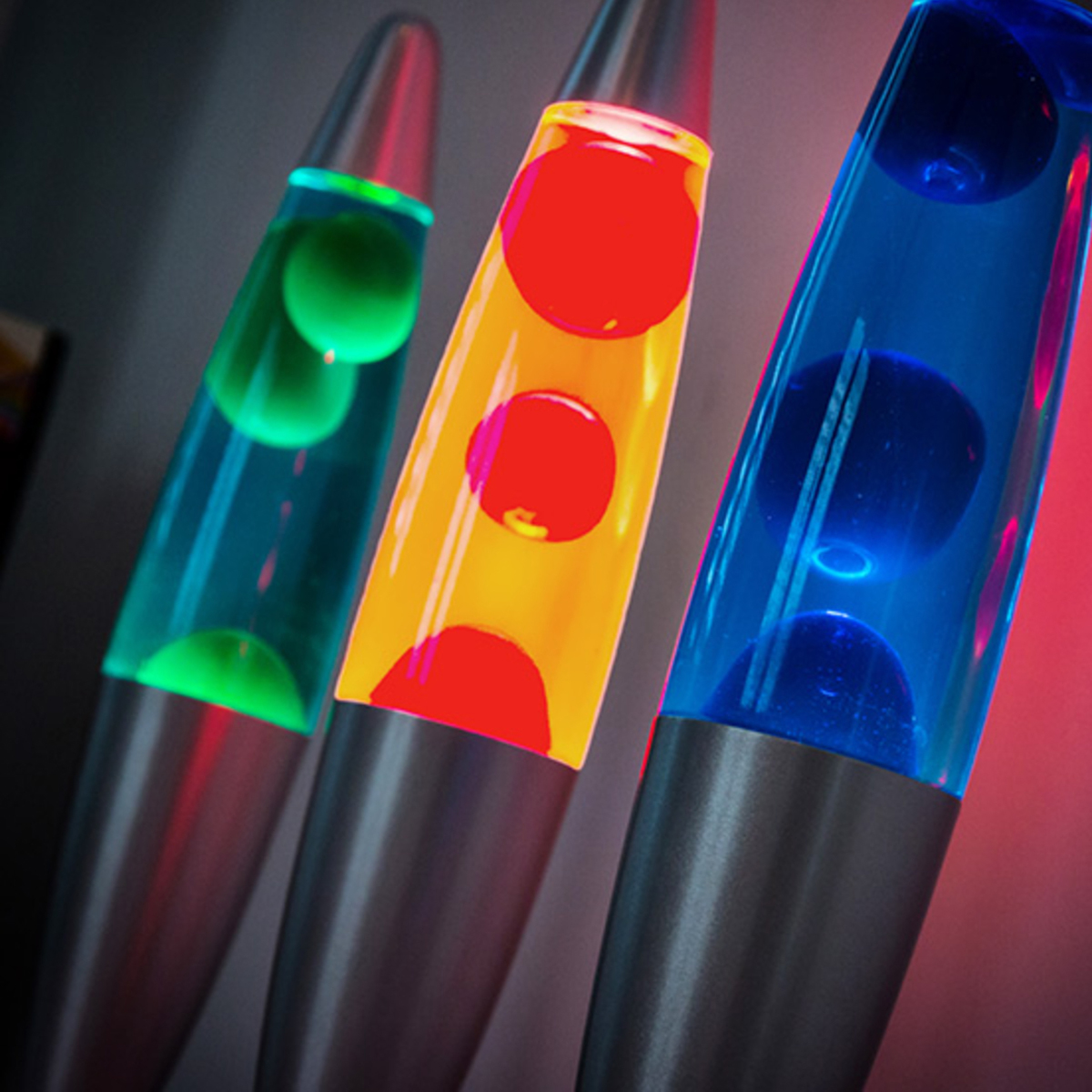 Tres lámparas de lava con lava y bases de diferentes colores. El fondo es oscuro, con luz roja y azul.