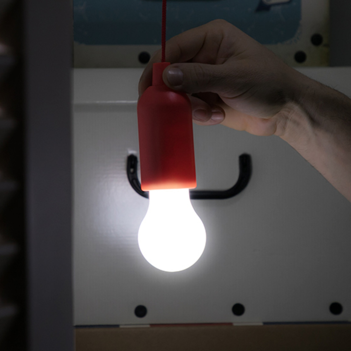 Mano sosteniendo lámpara roja en forma de botella con bombilla encendida. Fondo es un armario blanco.