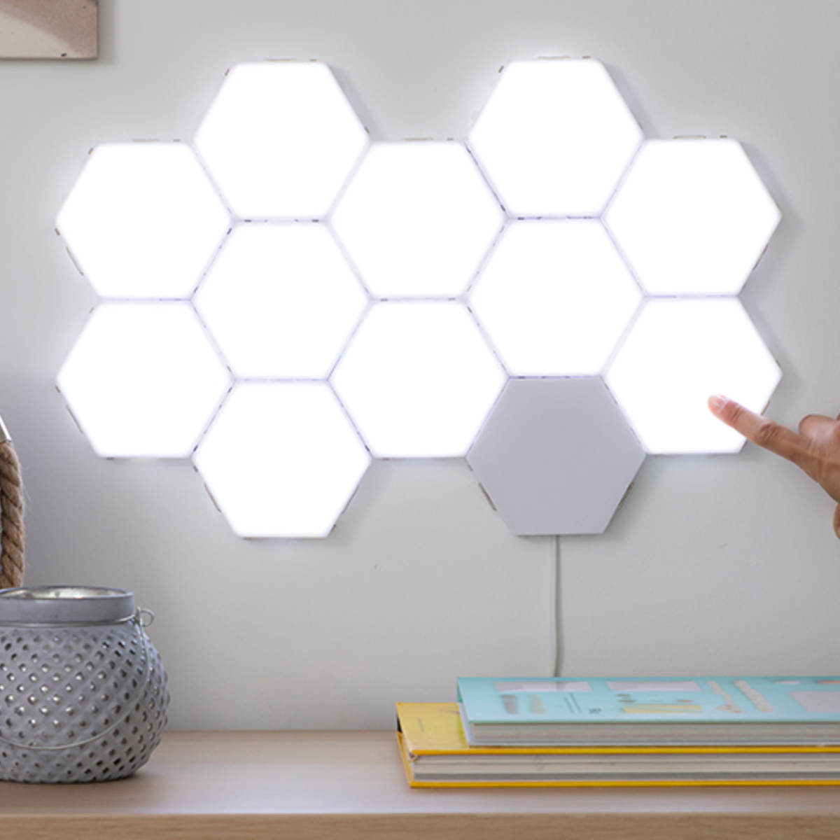 Una pared con un conjunto de luces hexagonales blancas, una mano tocando una. Hay un libro y una maceta en una mesa.