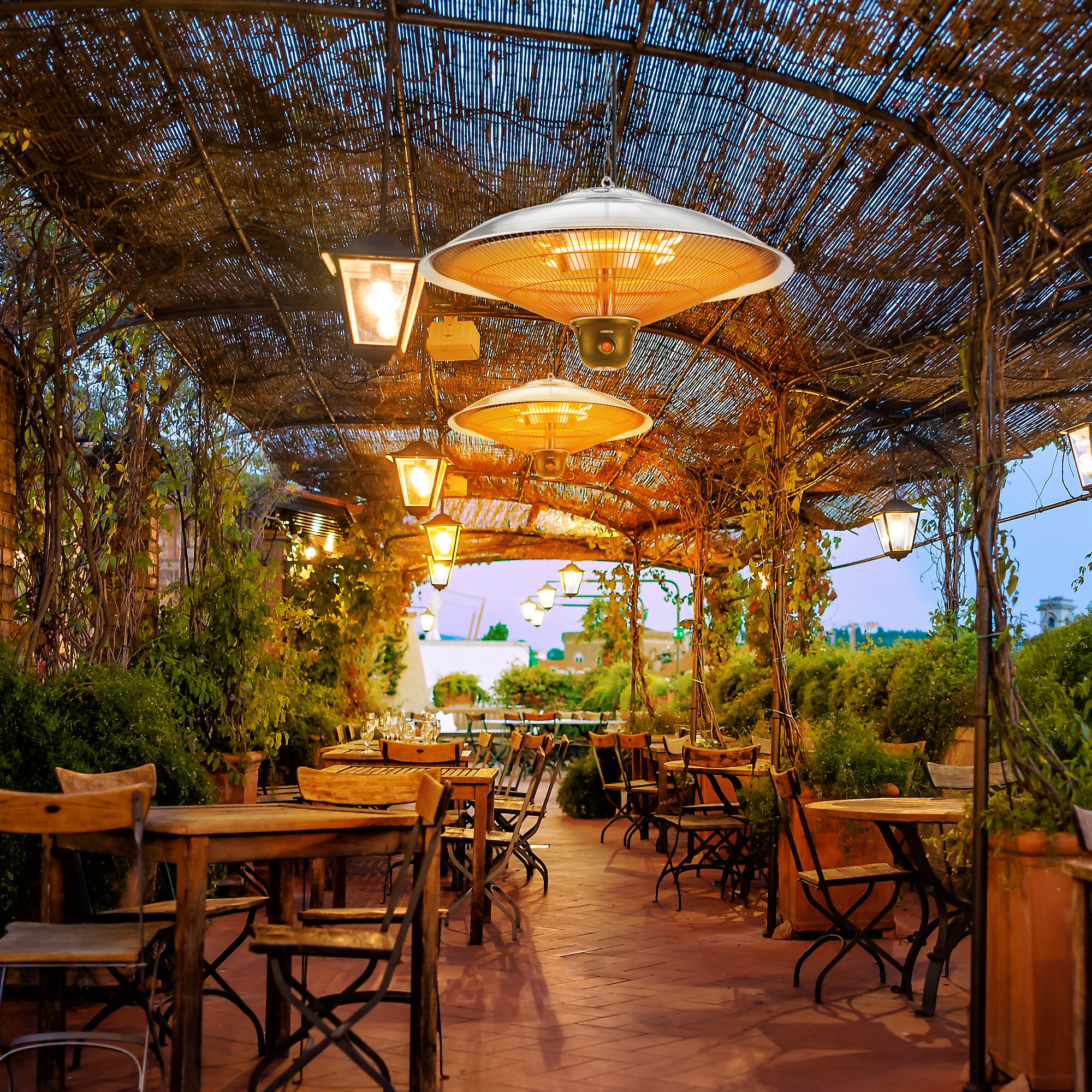 Außenrestaurant mit Tischen, Stühlen und Hängelampen unter einem gewebten Baldachin, mit Pflanzen und Stadtblick.