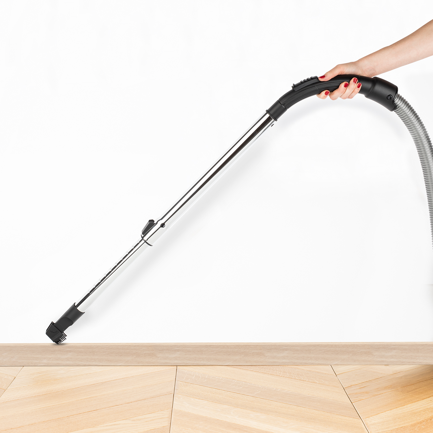 Dłoń trzymająca uchwyt odkurzacza, czyszcząca drewnianą podłogę przy białej ścianie.