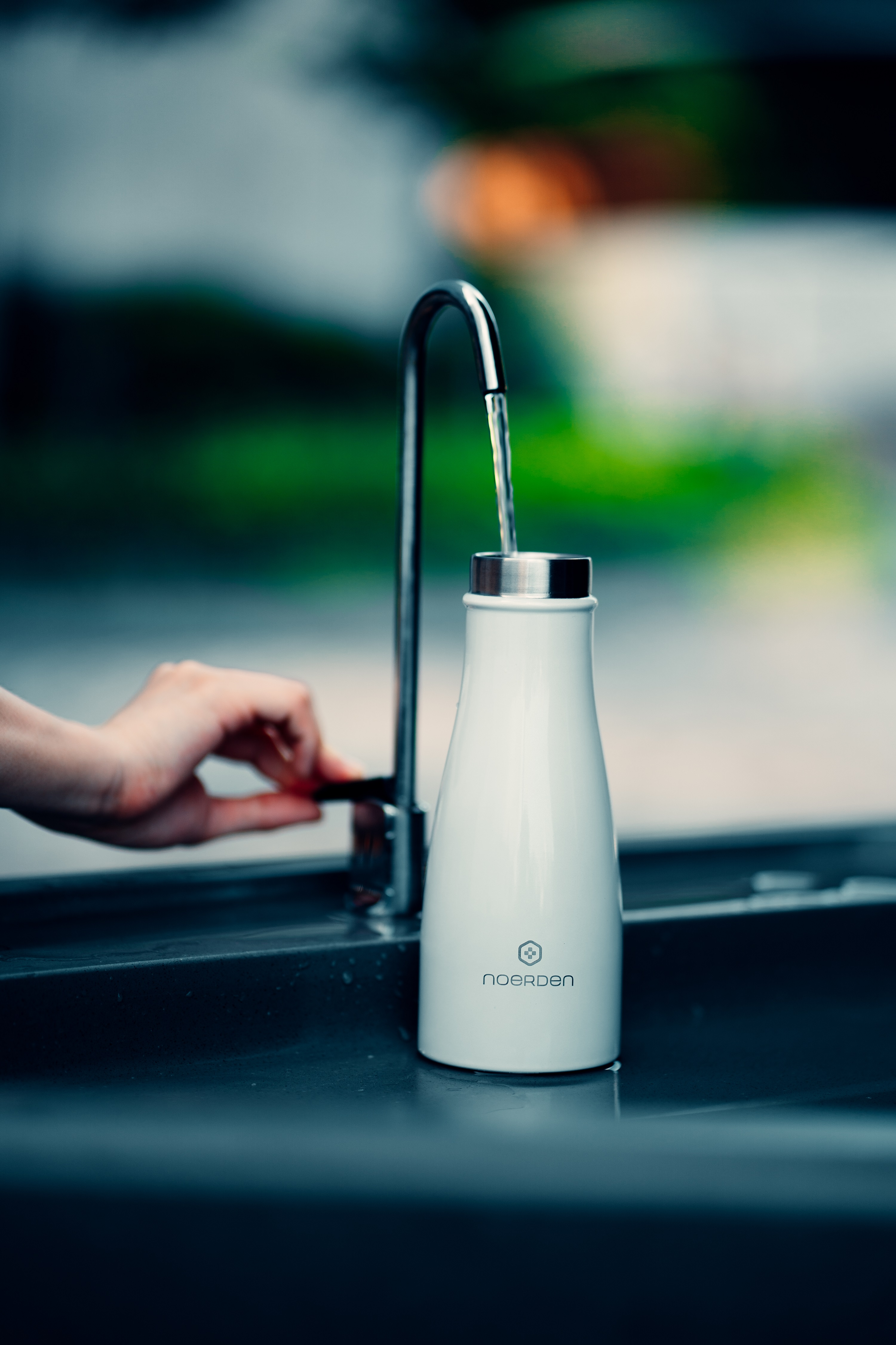 Weiße Wasserflasche, die an einem Wasserhahn gefüllt wird. Eine Hand dreht den Wasserhahn. Der Hintergrund ist unscharf.