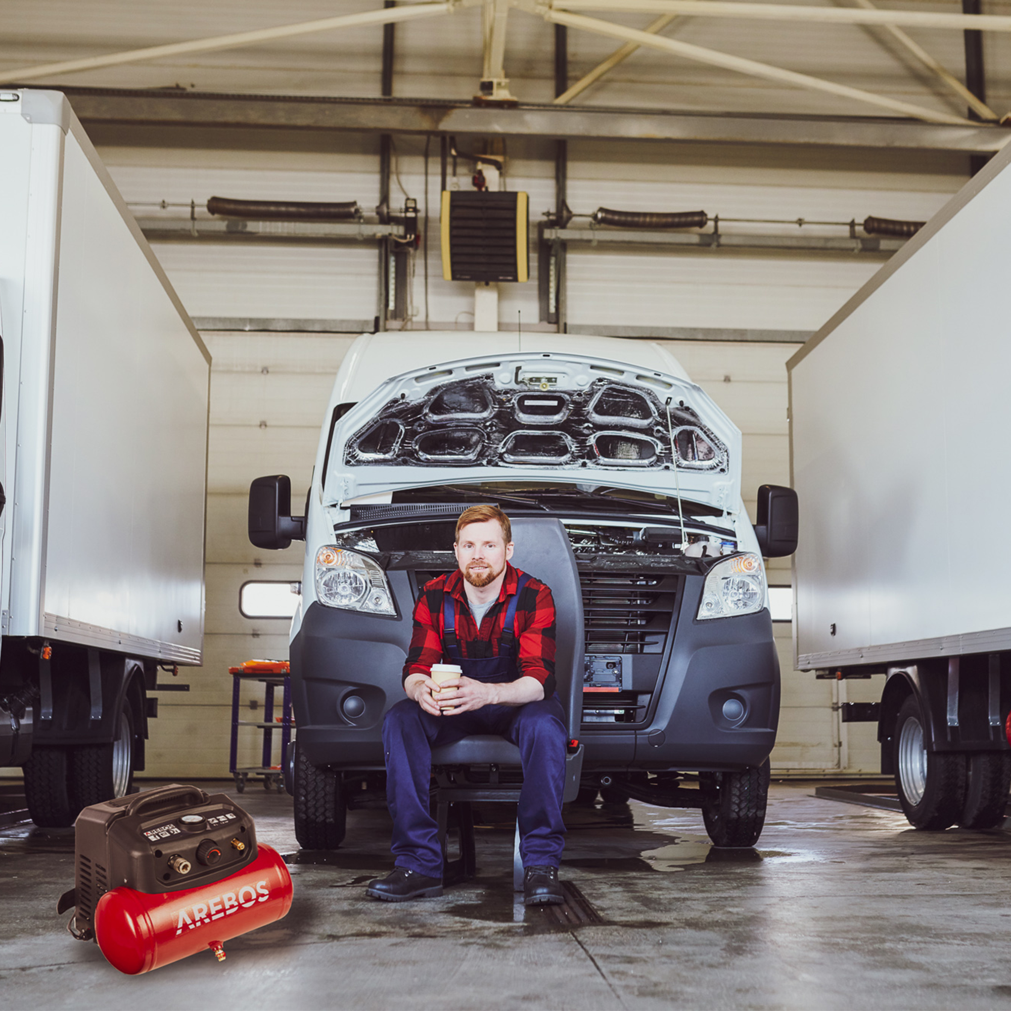 Mechaniker sitzt, hält Kaffee, in Garage mit LKW und rotem Luftkompressor.