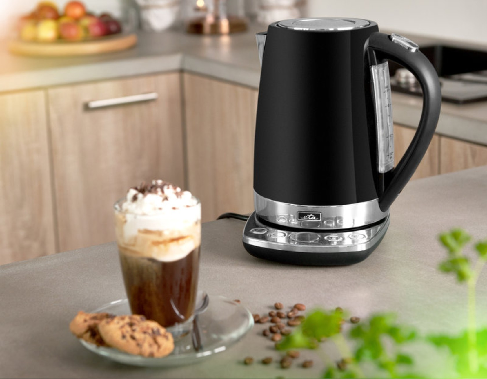 Schwarzer elektrischer Wasserkocher auf einer Theke. Ein Glas Kaffee mit Sahne und Keksen im Vordergrund.
