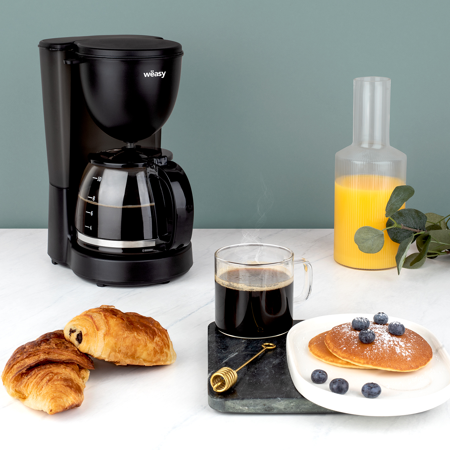 Schwarze Kaffeemaschine, Kaffee, Pfannkuchen, Croissant, Orangensaft auf einem Tisch. Der Hintergrund ist grün.