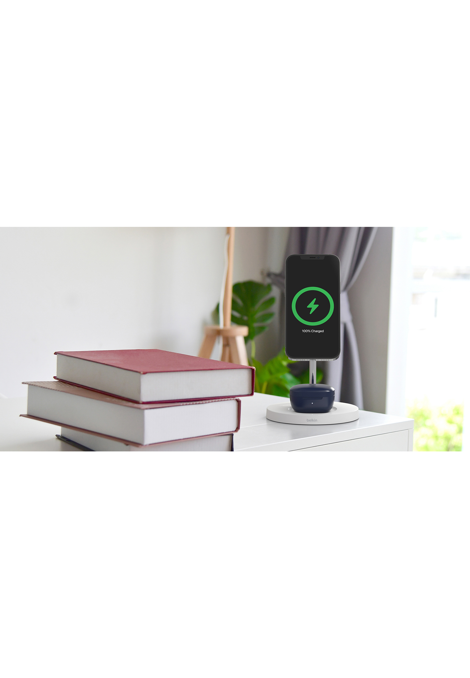 Uno smartphone in carica su una superficie bianca con libri e una lampada. Il telefono mostra 100% carica.