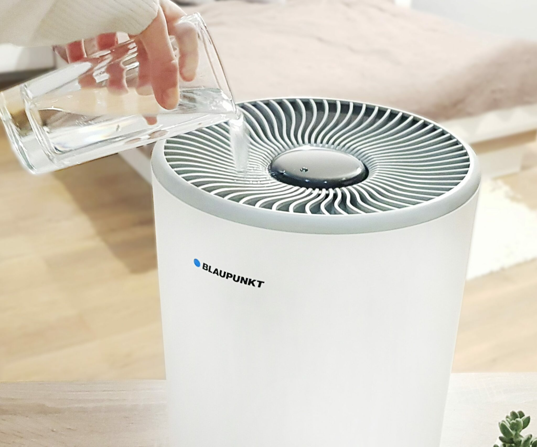 Una persona versa acqua in un purificatore d'aria bianco con logo blu, su un tavolo di legno.