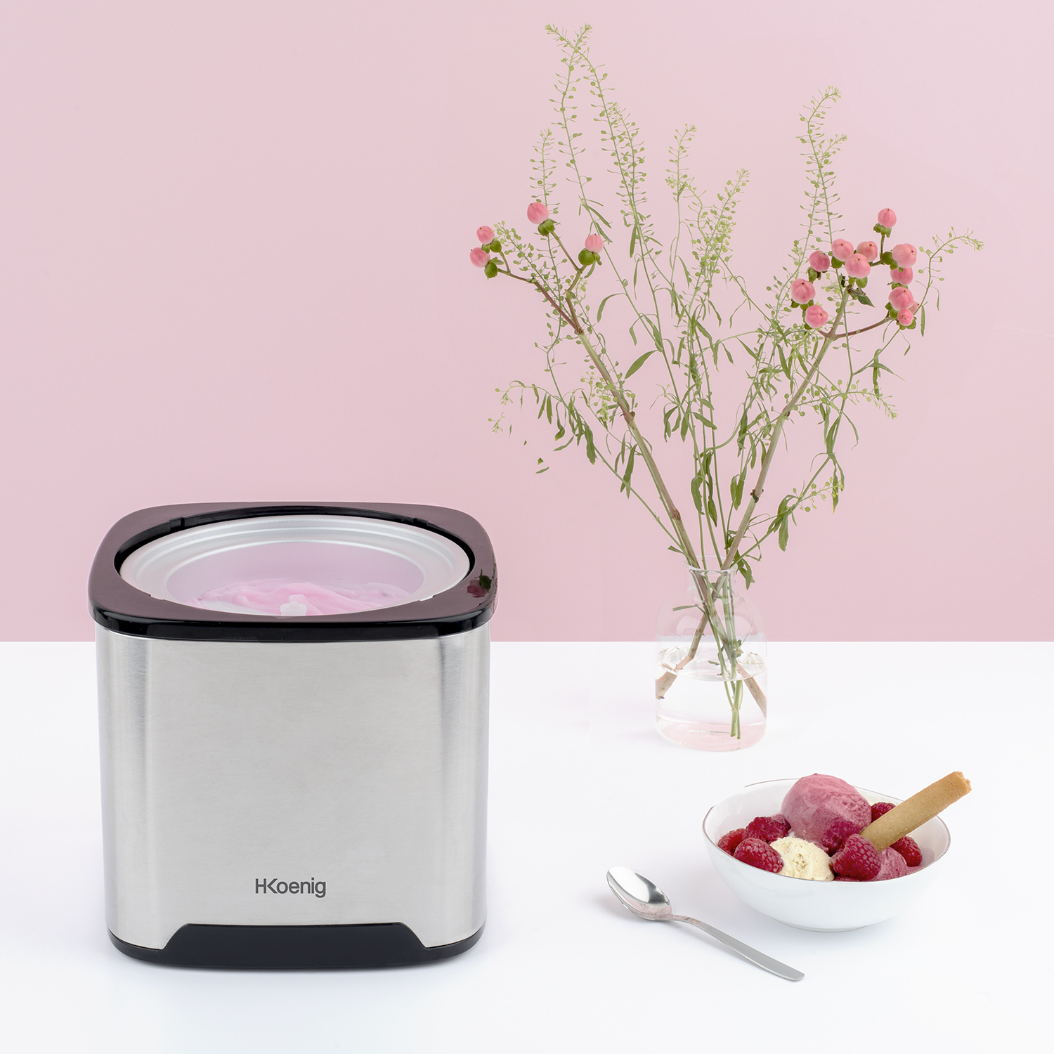 Maszyna do lodów i miska lodów z łyżką, malinami i wazonem z kwiatami.
