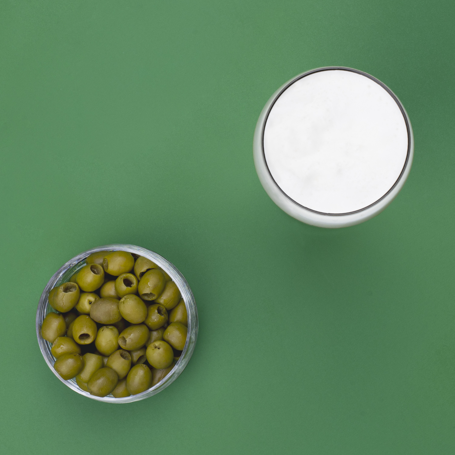 Bol de vidrio con aceitunas verdes, vaso de líquido sobre superficie verde.