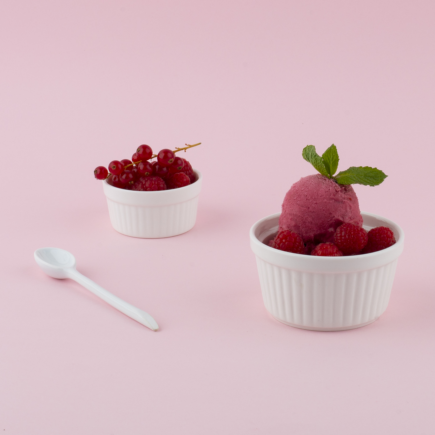 Zwei weiße Schalen mit roten Beeren und Eiscreme mit Minze auf rosa Hintergrund.
