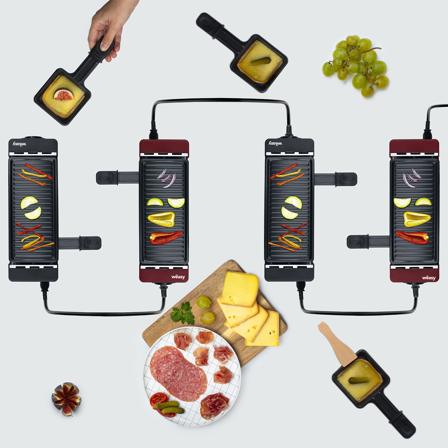 Ein Raclette-Grill-Setup mit verschiedenen Zutaten: Käse, Trauben, Salami und Gemüse. Weißer Hintergrund.