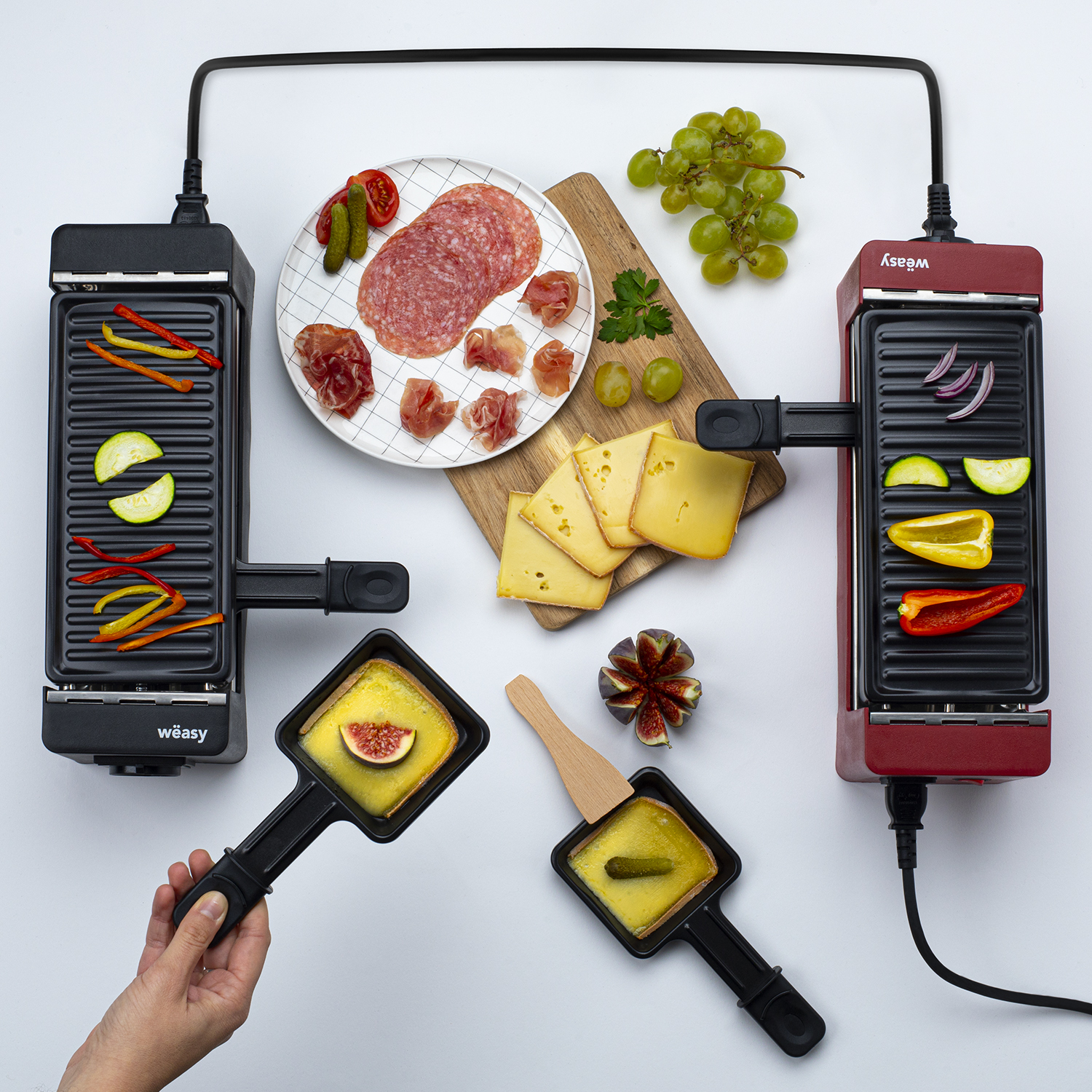 Raclette-Set mit zwei Grills, Lebensmittel wie Gemüse, Käse, Salami, Trauben und Feigen.