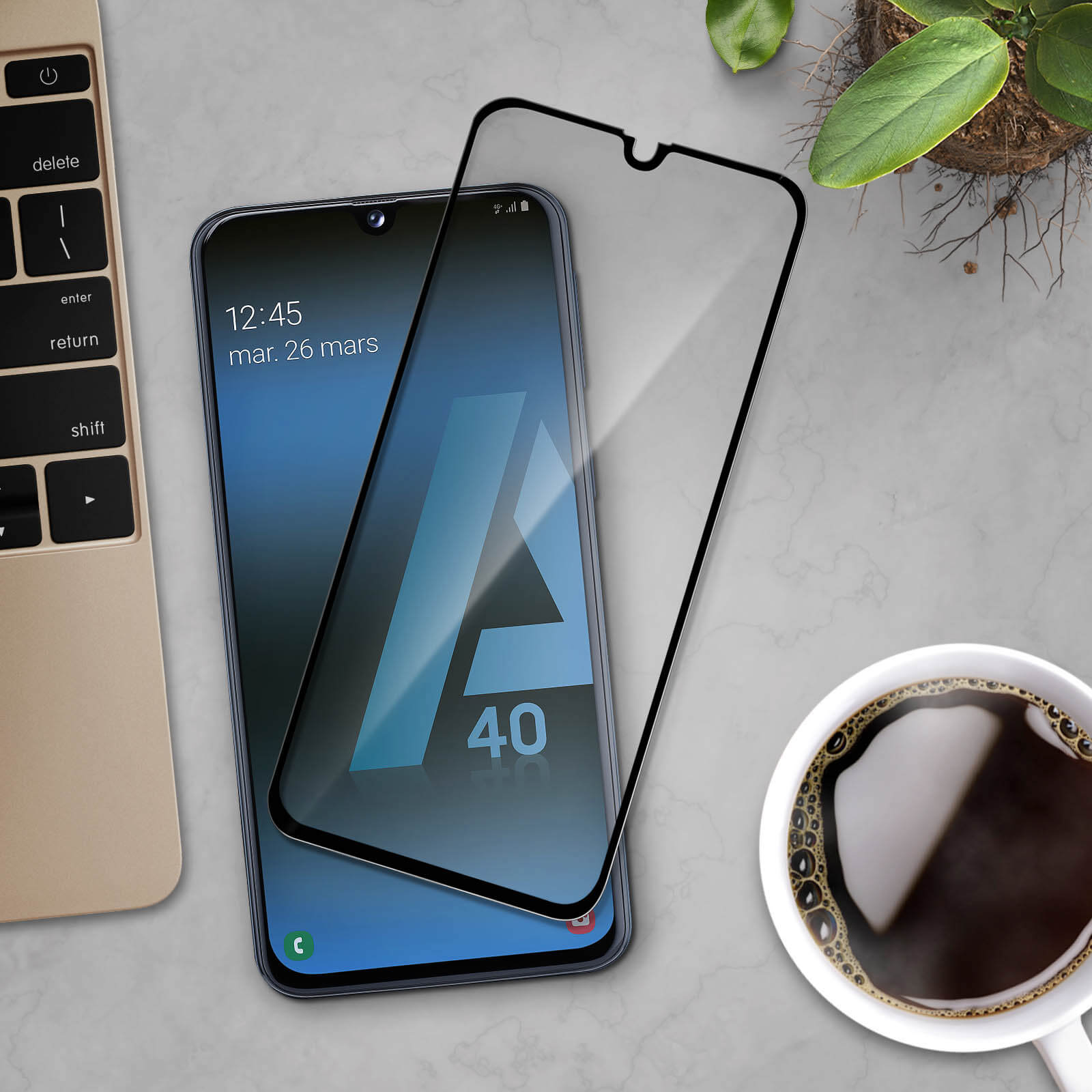 Smartphone con protezione schermo e una tazza di caffè, una pianta e una tastiera. Superficie grigia.