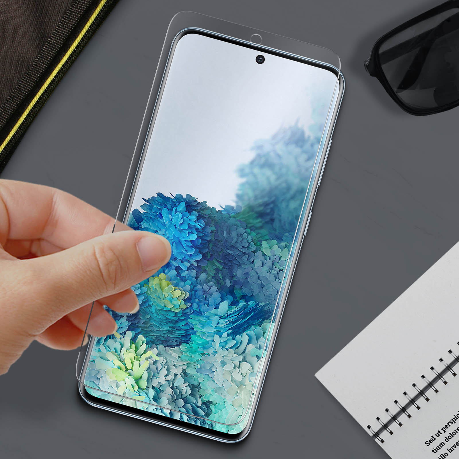 Mano applica protezione schermo al telefono. Sfondo floreale. Occhiali da sole e blocco note su superficie grigia.
