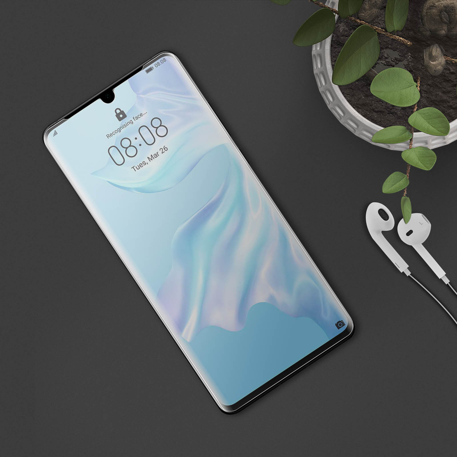 Telefono su superficie scura con auricolari e pianta sullo sfondo.