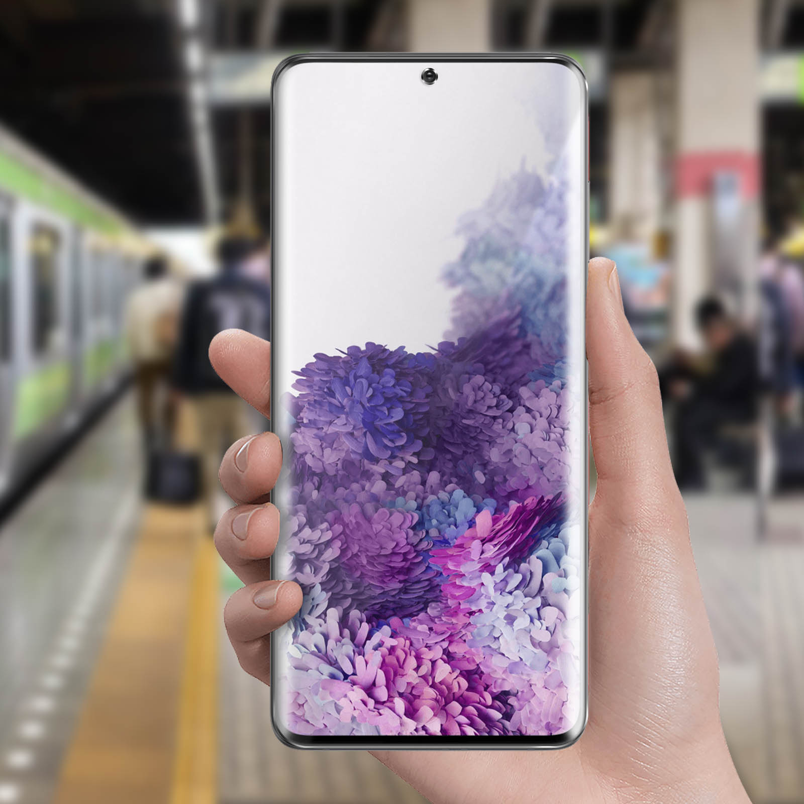 Una mano tiene un telefono che mostra fiori viola. Lo sfondo sfocato mostra una stazione ferroviaria con persone e un treno.