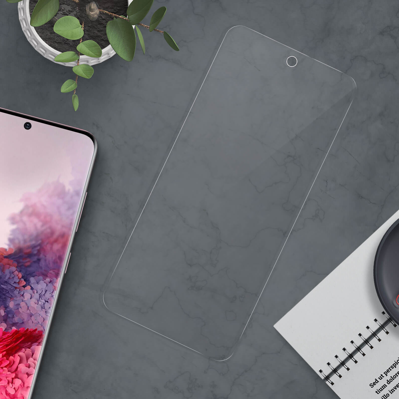Uno smartphone con una protezione per lo schermo su una superficie grigia. Sono presenti anche una piccola pianta e un taccuino.