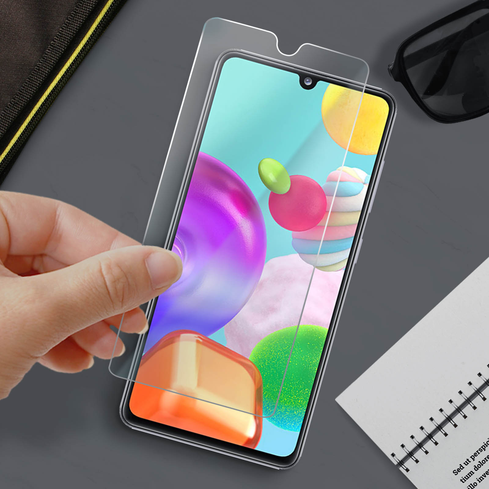 Mano tiene una protezione schermo sopra un telefono. Il telefono mostra forme astratte colorate. Occhiali da sole e blocco note presenti.