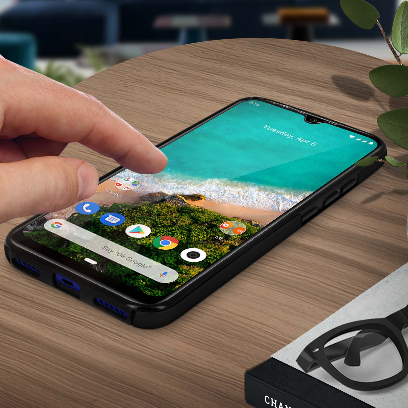 Main touchant l'écran du téléphone. Téléphone sur la table. L'écran affiche une image de plage. Lunettes à droite.