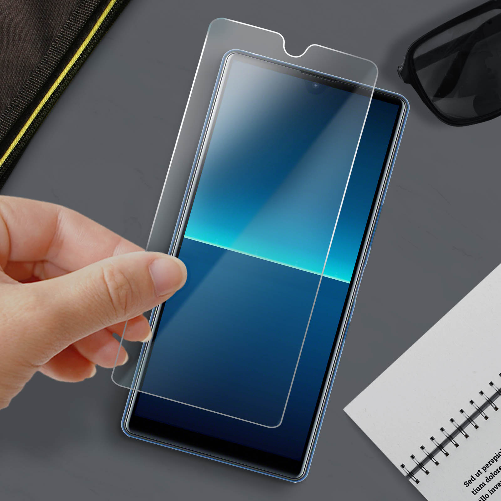 Una mano che tiene una protezione per lo schermo su uno smartphone blu. Sono presenti anche occhiali da sole, un blocco note e una borsa.