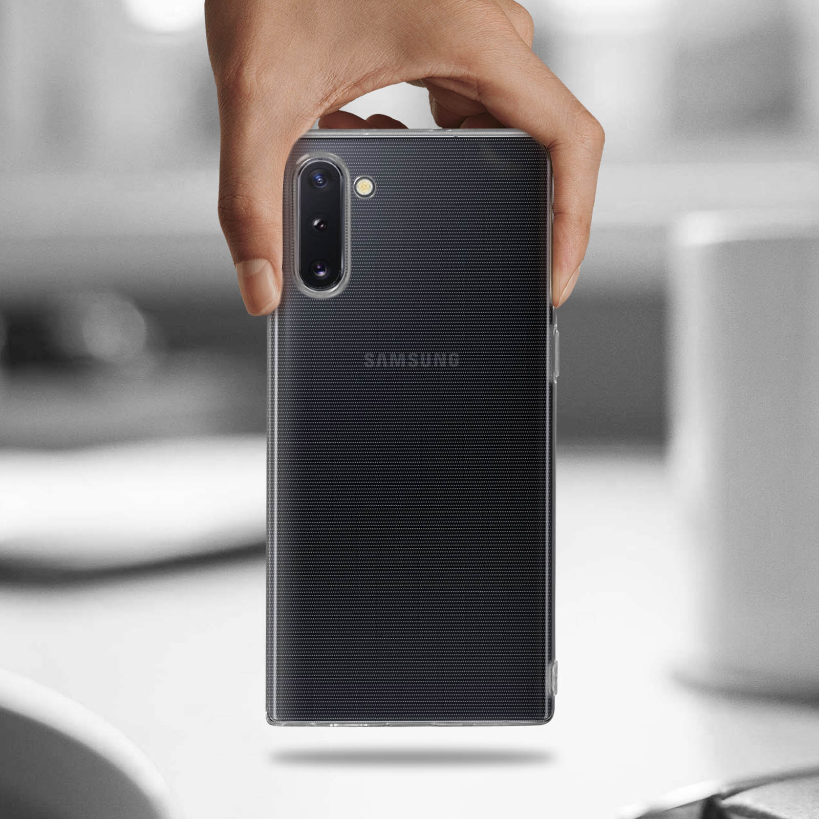 Mano che tiene un telefono nero in una custodia trasparente. Lo sfondo è sfocato e grigio.