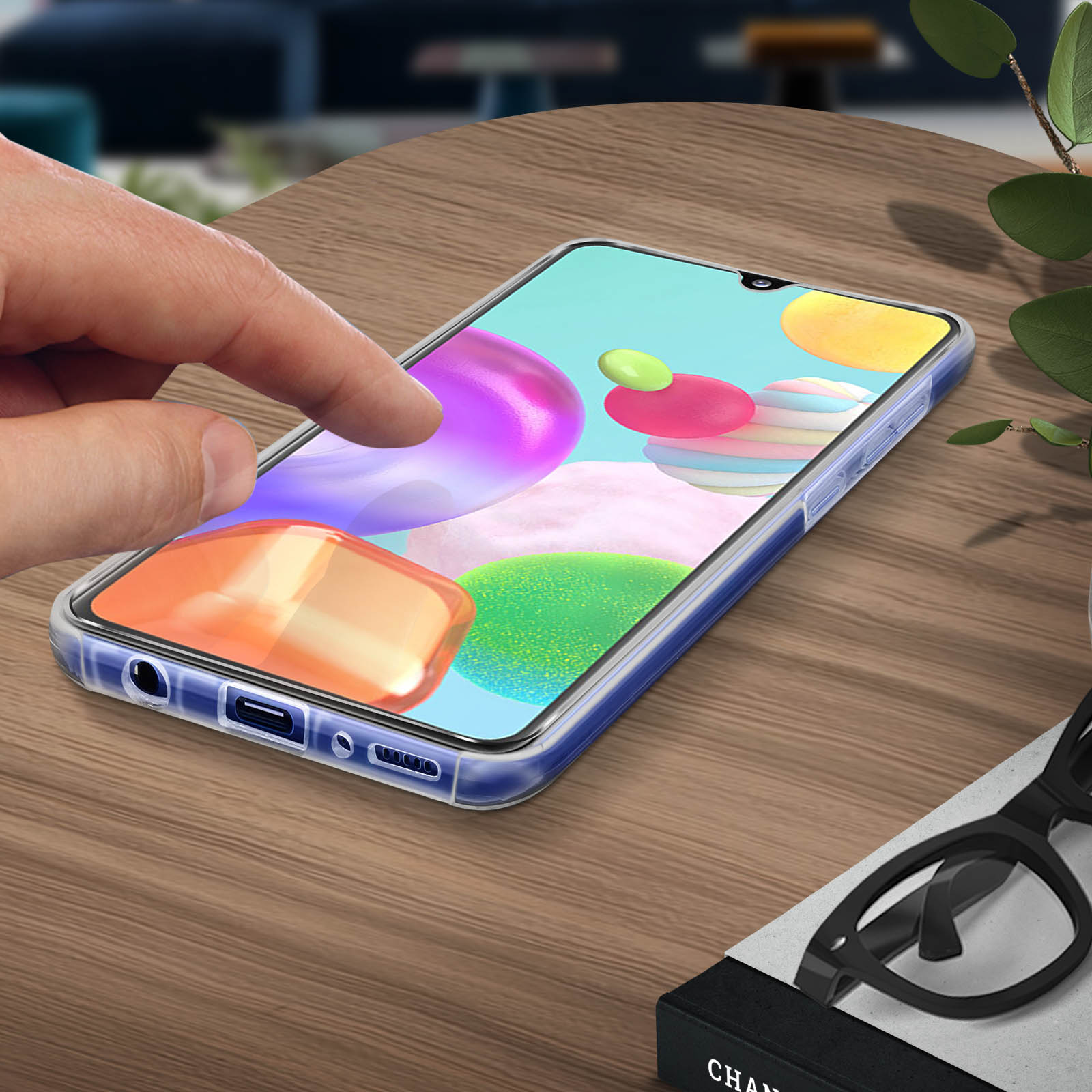 Een hand raakt het telefoonscherm aan met kleurrijke pictogrammen. Plant en bril op de achtergrond.