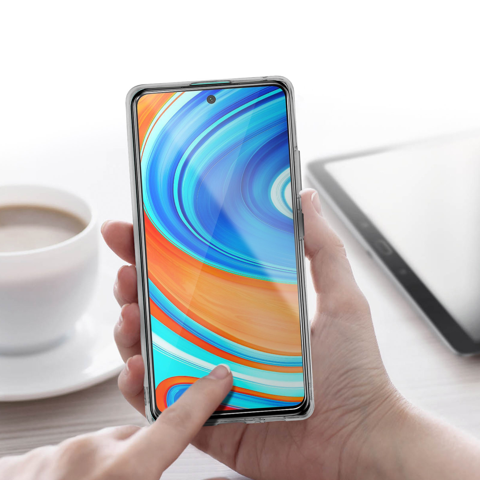 Eine Hand berührt ein Telefon mit Displayschutz. Im Hintergrund befinden sich eine Tasse und ein Tablet.
