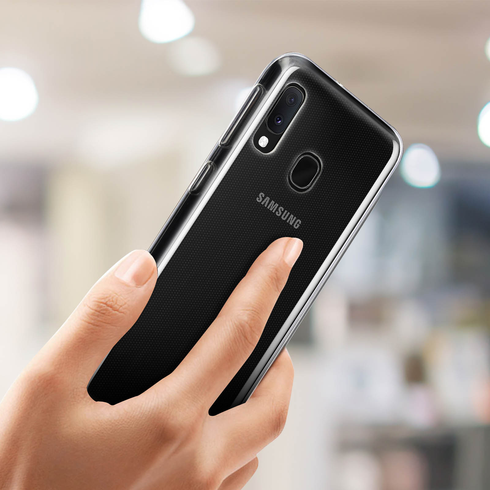Una mano che tiene un telefono Samsung nero, con il logo Samsung visibile. Lo sfondo è sfocato.